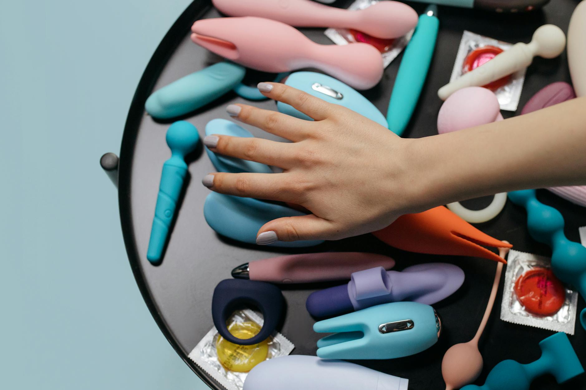 A hand reaching toward a collection of colorful adult toys on a neutral surface